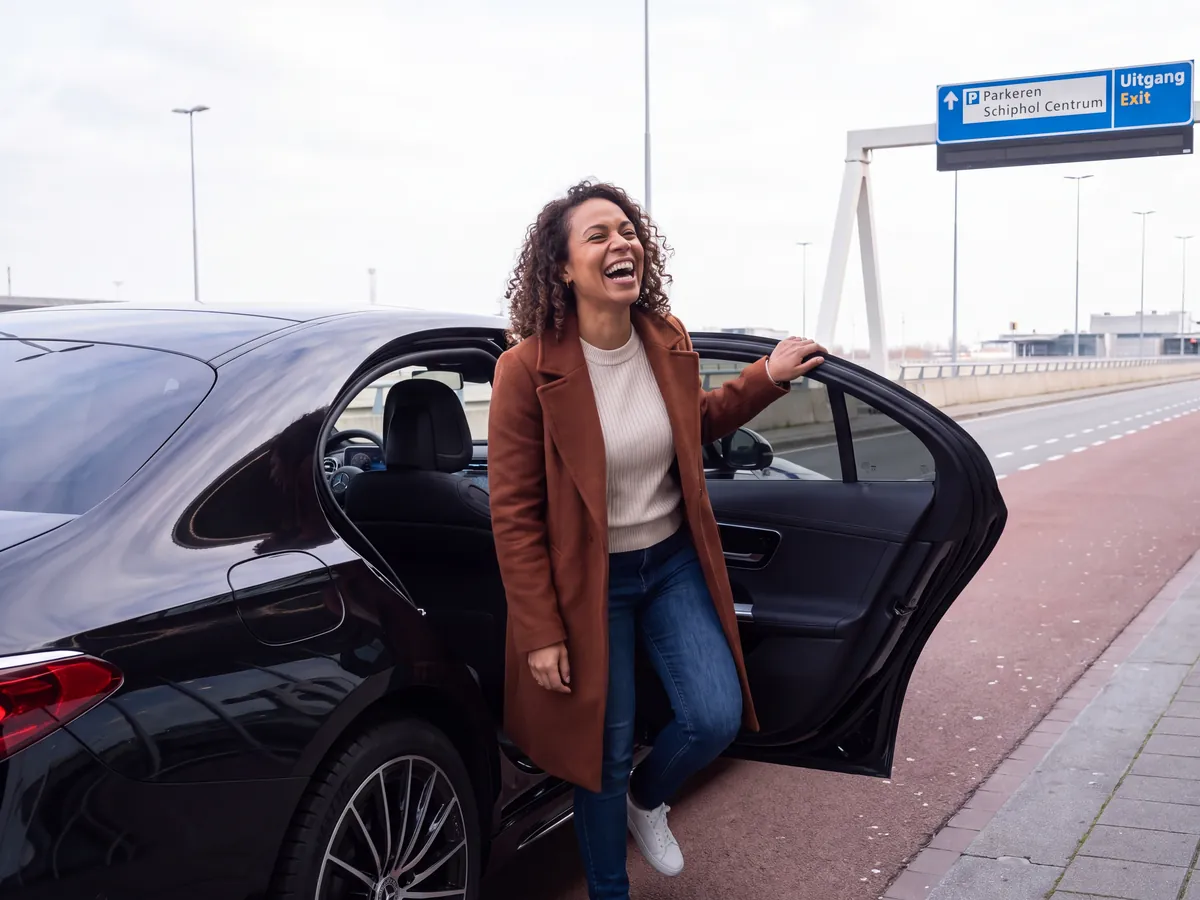 Smiling woman in a brown coat stepping out of a black sedan at Schiphol