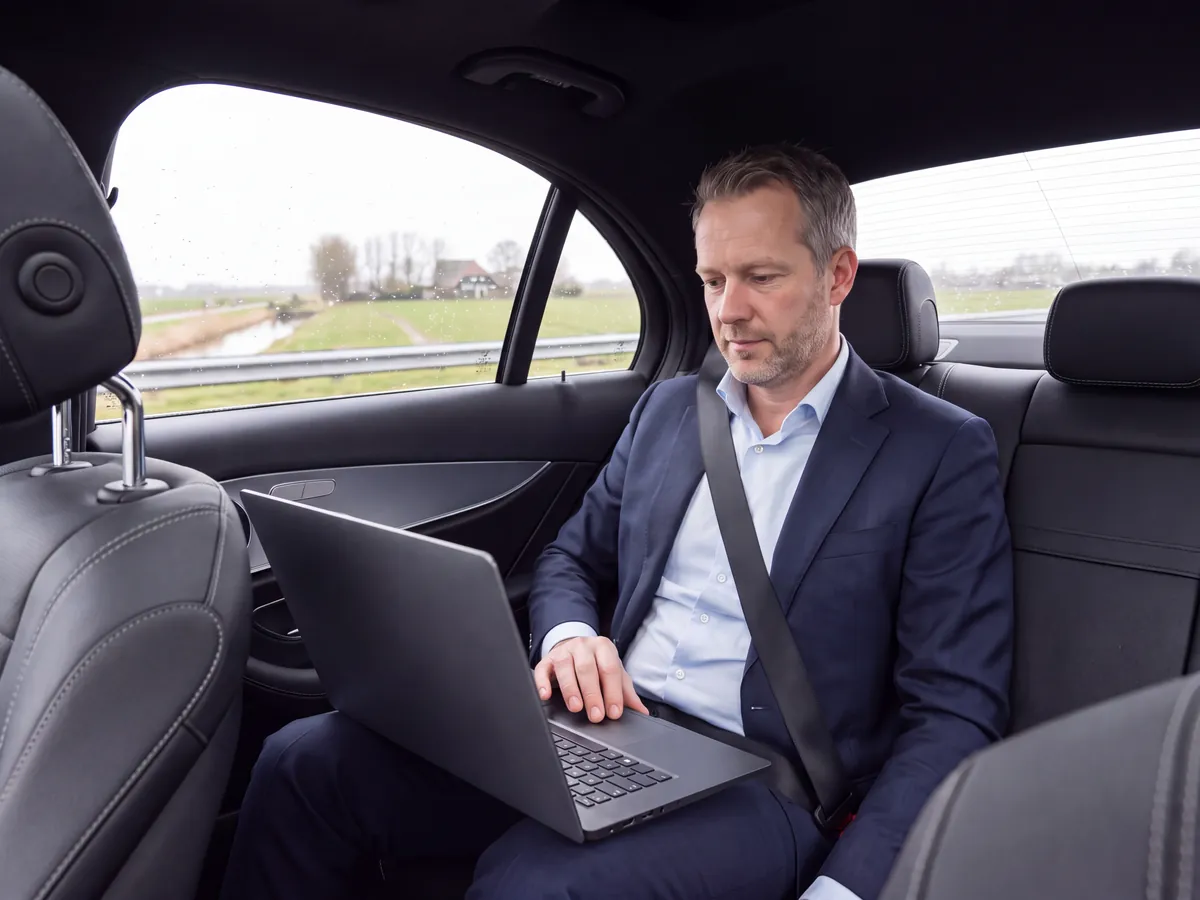 Man in a suit working on a laptop in the back seat of a car, with fields outside
